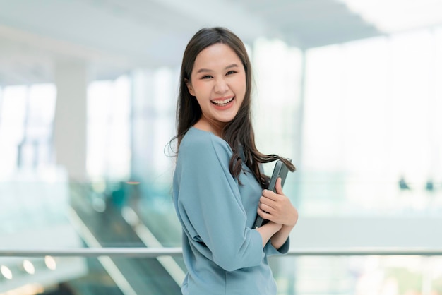 Asian sorridente allegro femmina nomade digitale mano tenere tablet dispositivo guardare la fotocamera ritratto shotfelicità sorridente asia donna in piedi in ufficio college corridoio con positivo atteggiamento sorridente