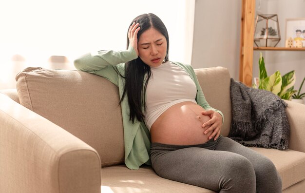 Asiatica giovane donna incinta che soffre di mal di pancia seduta su un divano nel soggiorno di casa