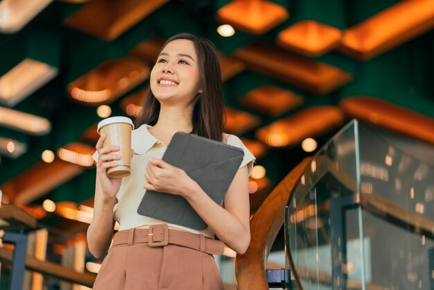 asiatico femminile creativo freelance digitale nomade tenere in mano tablet e tazza di caffè sorridente allegro casual rilassarsi camminando nel bar ristorante downshifting stile di vita giovane donna millenaria nella caffetteria