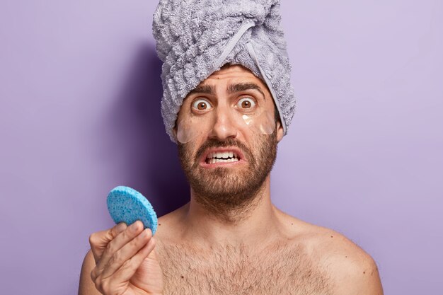 Aspetto maschile nervoso con espressione del viso preoccupata e dispiaciuta, trattiene la spugna cosmetica, applica cerotti idrogel per rimuovere le borse sotto gli occhi