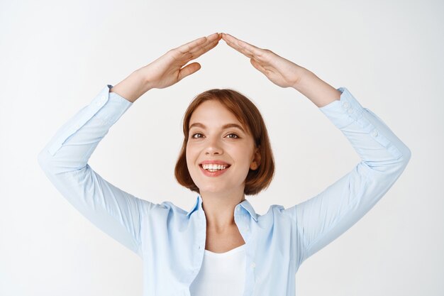 Assicurazione e concetto immobiliare. Donna sorridente in camicetta che fa il tetto della mano sulla testa, mostrando il gesto di casa, in piedi sul muro bianco