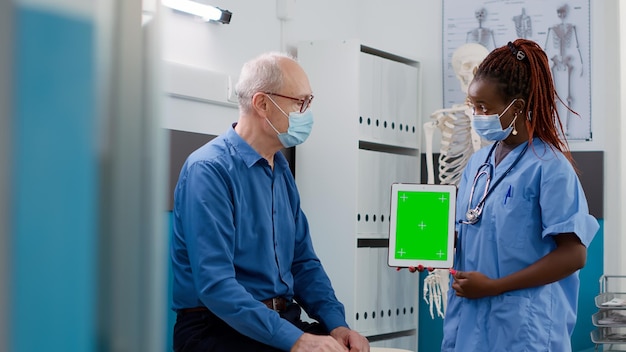 Assistente che mostra tablet con schermo verde al paziente anziano durante la pandemia covid 19. Esaminando il modello di mockup isolato con copyspace vuoto e display chromakey durante la visita di controllo.