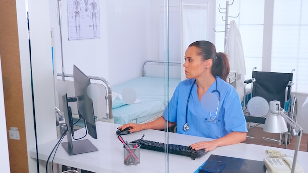 Assistente medico digitando sul computer con un monitor seduto dietro una parete di vetro in ufficio. Medico in medicina uniforme per la scrittura di un elenco di pazienti consultati e diagnosticati, che effettuano ricerche.