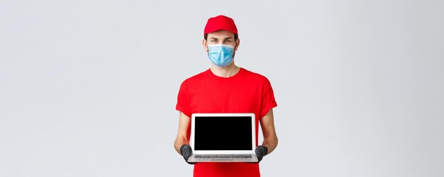 Assistenza clienti covid19 pacchetti di consegna ordini online concetto di elaborazione Corriere sorridente in uniforme rossa maschera facciale e guanti che mostrano la pagina web dello schermo del laptop dell'azienda