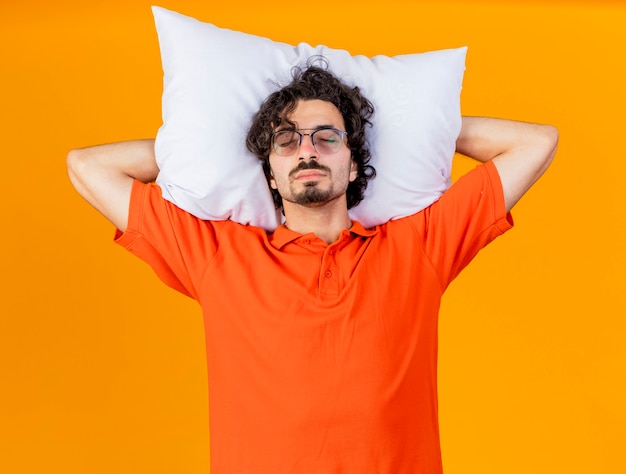 Assonnato giovane uomo malato caucasico con gli occhiali che tengono cuscino sotto la testa fingere di dormire con gli occhi chiusi isolati sulla parete arancione