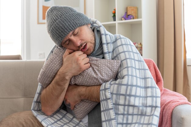 Assonnato uomo malato con sciarpa intorno al collo indossando cappello invernale avvolto in plaid tenendo e mettendo la testa sul cuscino seduto sul divano in soggiorno