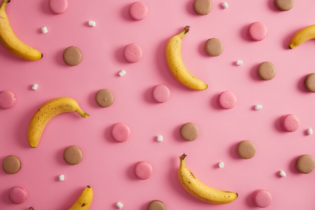 Assortimento di dolci deliziosi amaretti francesi colorati, marshmallow bianco e banane su sfondo rosa. Biscotti ipercalorici malsani e frutta tropicale sana. Idea per il tuo dessert