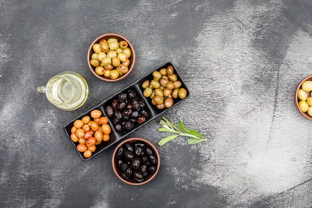 Assortimento di olive fredde in un piatto nero e ciotole di argilla con un barattolo di olio d'oliva e foglie di ulivo vista dall'alto su grigio scuro grunge