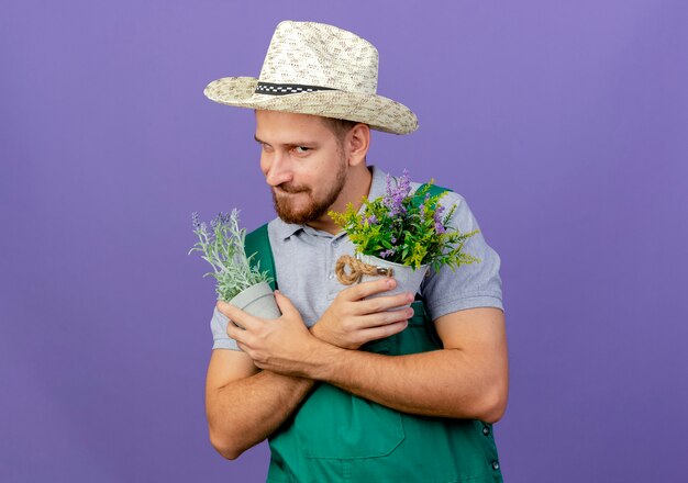 Astuto giovane giardiniere slavo bello in uniforme e cappello in possesso di vasi da fiori tenendo le mani incrociate e labbro mordace isolato sul muro viola con spazio di copia