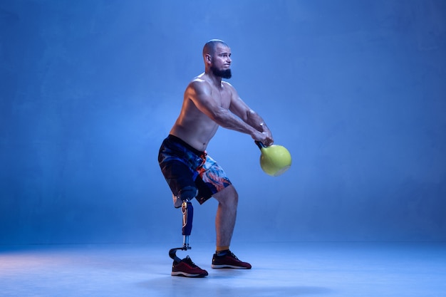 Atleta con disabilità o amputato isolato sulla parete blu. Sportivo maschio professionista con allenamento di protesi di gamba con pesi al neon. Sport per disabili e superamento, concetto di benessere.