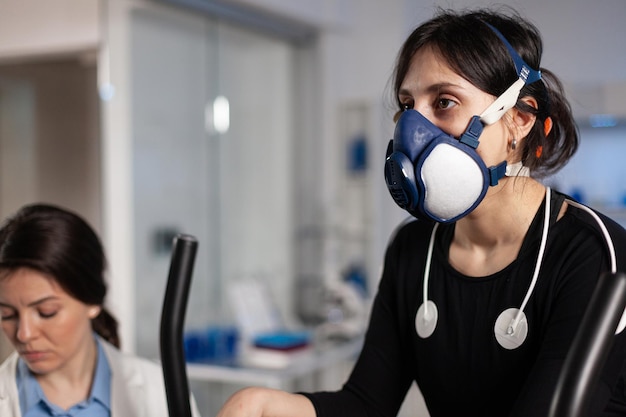 Atleta donna con maschera in esecuzione su fitness bycle training resistenza del corpo mentre medico specialista che misura la frequenza cardiaca monitorando i dati EGK in laboratorio. Sportiva con elettrodi medici su di esso