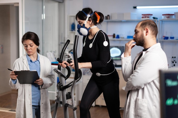 Atleta donna con maschera in esecuzione su palestra bycle training resistenza del corpo mentre medico ricercatore che misura la frequenza cardiaca monitorando i dati EGK in laboratorio. Sportiva con elettrodi medici su di esso