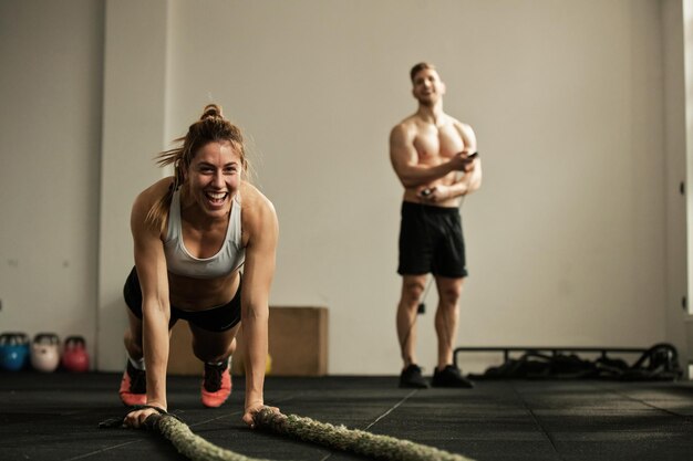 Atleta femminile allegra che fa flessioni mentre si esercita con la corda da battaglia e si diverte in una palestra C'è un uomo sullo sfondo