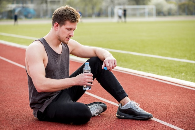 Atleta maschio che si siede sulla pista di corsa che giudica bottiglia di acqua disponibila