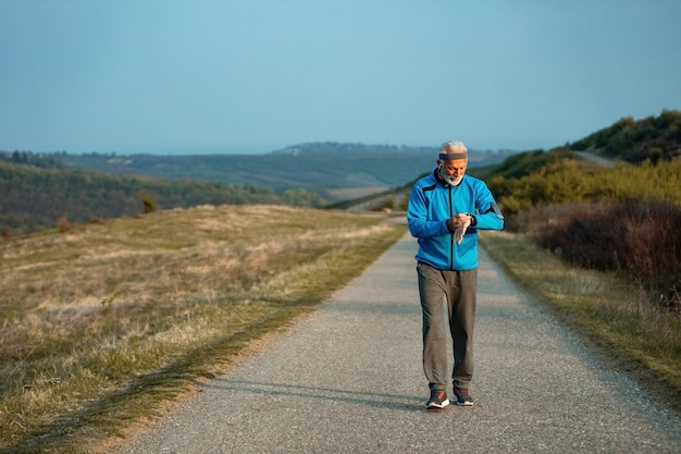 Atleta senior attivo che controlla la traccia del polso sull'orologio intelligente mentre cammina per strada in natura Spazio di copia