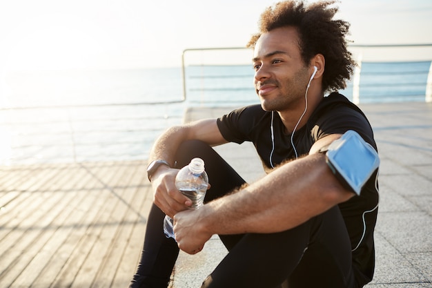 Atleta uomo dalla pelle scura in forma sorridente con capelli folti che beve acqua dalla bottiglia di plastica dopo un duro allenamento in riva al mare. Colpo di corridore maschio in top nero e leggings che riposa dopo il successo