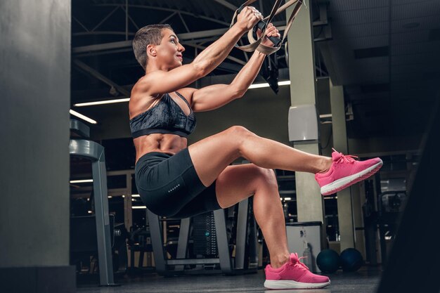 Atletica donna di mezza età con i capelli corti che fa allenamenti per le gambe con strisce di sospensione trx in un club di palestra.