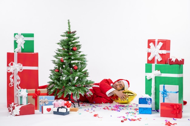 Atmosfera festosa vacanza con leepings babbo natale sdraiato dietro l'albero di natale vicino a regali in diversi colori su sfondo bianco stock photo