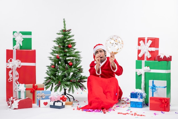 Atmosfera natalizia con Babbo Natale spaventato che tiene palloncino seduto vicino all'albero di Natale e regali in diversi colori su sfondo bianco
