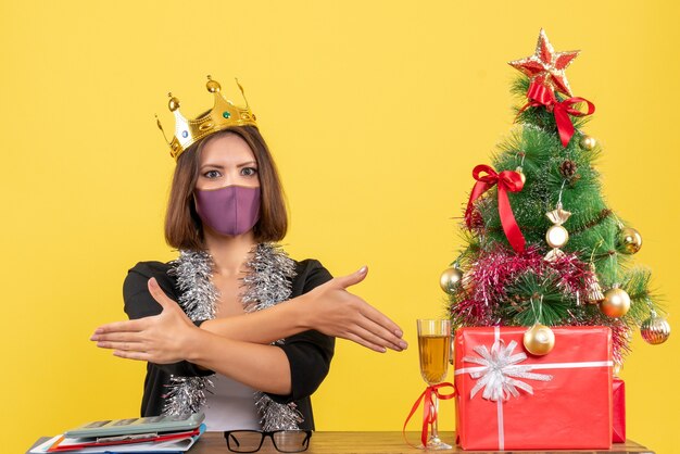 Atmosfera natalizia con bella signora in tuta con maschera medica e maschera da portare che accoglie i suoi ospiti in ufficio su giallo