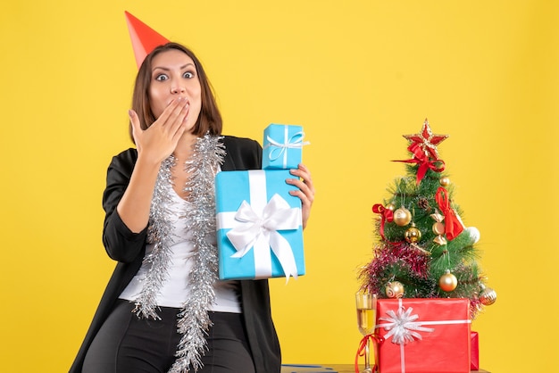 Atmosfera natalizia con bella signora sorpresa emotiva con il cappello di Natale che tiene i regali in ufficio su giallo