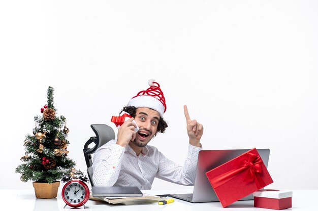 Atmosfera natalizia con giovane barbuto uomo d'affari con cappello di Babbo Natale alzando il suo regalo e indicando sopra su sfondo bianco