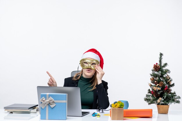 Atmosfera natalizia con giovane donna con cappello di Babbo Natale e maschera da portare seduto a un tavolo su sfondo bianco