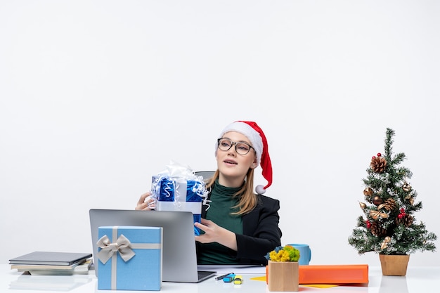 Atmosfera natalizia con giovane donna con cappello di Babbo Natale e occhiali da vista seduto a un tavolo che riceve un regalo su priorità bassa bianca