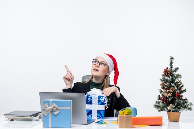 Atmosfera natalizia con giovane donna con cappello di Babbo Natale e occhiali da vista seduto a un tavolo con regalo e mostrando sopra su sfondo bianco