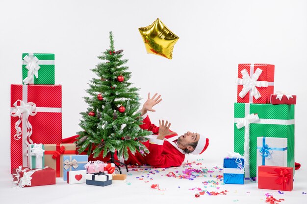 Atmosfera natalizia con il giovane Babbo Natale sdraiato dietro l'albero di Natale vicino a doni in diversi colori su sfondo bianco stock photo