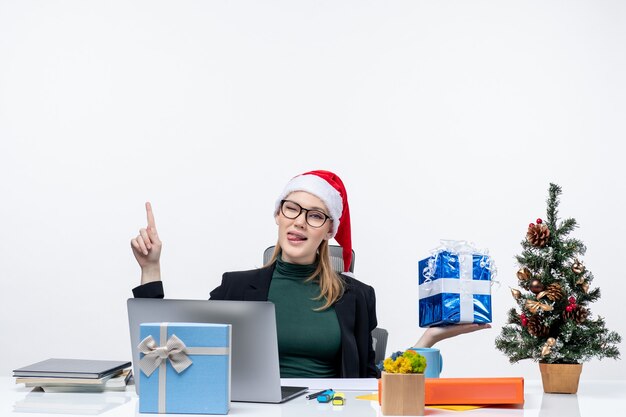 Atmosfera natalizia con positivo divertente giovane donna con cappello di babbo natale e indossando occhiali seduto a un tavolo che mostra il dono rivolto sopra su sfondo bianco