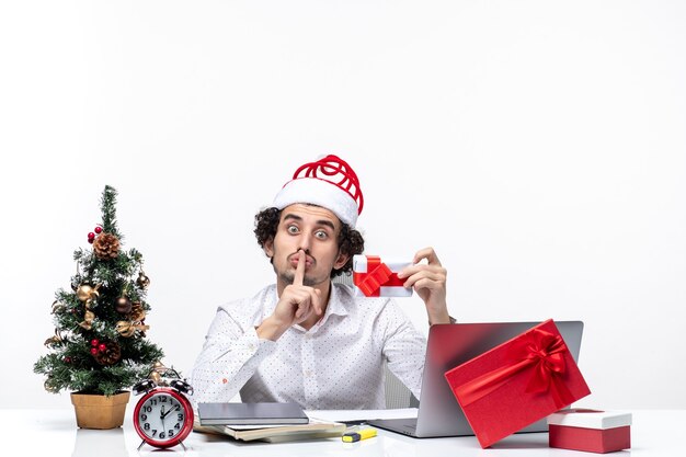 Atmosfera natalizia con uomo d'affari sorpreso con cappello di Babbo Natale alzando il suo regalo e facendo gesto di silenzio su priorità bassa bianca