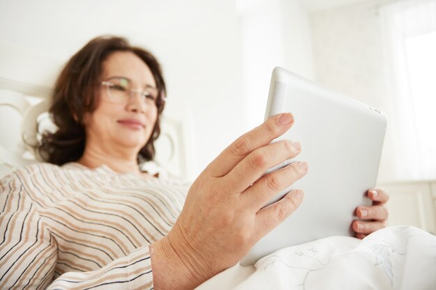 Attenta e allegra donna matura utilizzando un tablet pc sdraiata sul suo letto in una camera da letto