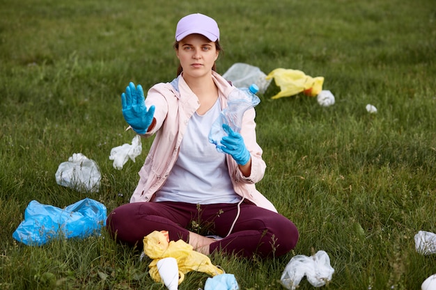 Attivista che raccoglie immondizia nel campo, sedendosi sull'erba verde e tenendo bottiglia di plastica