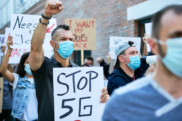 Attivista maschio con il pugno alzato che protesta contro la rete 5G mentre indossa una maschera protettiva durante le manifestazioni pubbliche