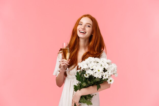 Attraente, allegra ragazza di b-day con i capelli rossi ricci, ridendo e guardando la telecamera spensierata mentre parla con gli amici durante la festa, la festa di compleanno, tenendo in mano un bouquet di fiori bianchi e un bicchiere di champagne.