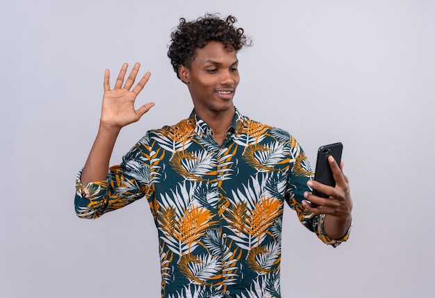 Attraente bell'uomo di carnagione scura con capelli ricci in camicia stampata foglie parlando con un amico durante la videochiamata e agitando la mano sulla fotocamera anteriore del telefono cellulare