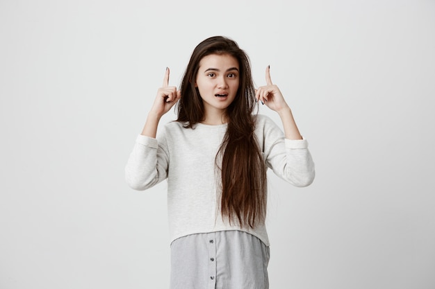 attraente bella donna eccitata con i capelli lunghi neri che punta le dita verso l'alto con sguardo sorpreso, con espressione allegra e felice del viso