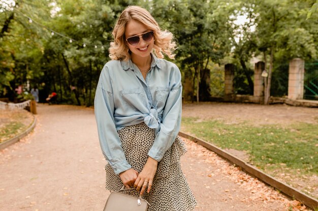 Attraente bionda candida donna che cammina nel parco in abito elegante indossando occhiali da sole eleganti e borsa