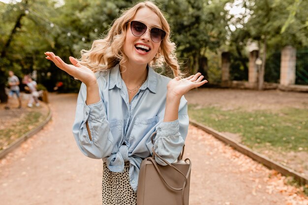 Attraente bionda candida donna che cammina nel parco in abito elegante indossando occhiali da sole eleganti e borsa