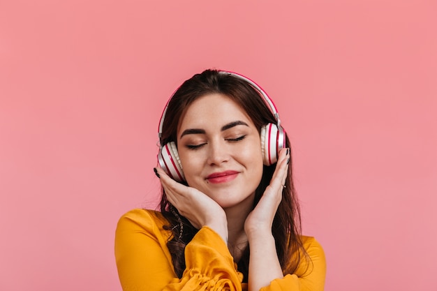 Attraente bruna con il sorriso e gli occhi chiusi ascoltando la canzone in cuffia. Signora in camicetta giallo brillante in posa sulla parete isolata.