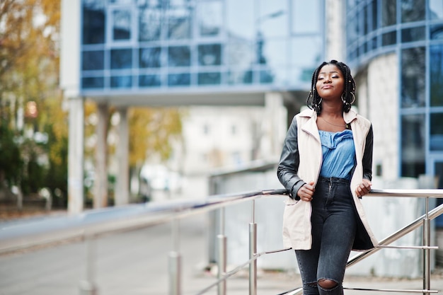 Attraente donna afroamericana con dreadlocks in giacca posata vicino alle ringhiere contro un moderno edificio a più piani