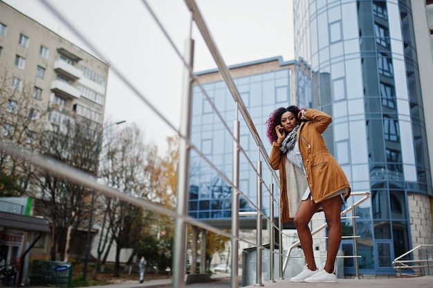 Attraente donna afroamericana riccia in cappotto marrone posata vicino alle ringhiere contro un moderno edificio a più piani che parla sul telefono cellulare