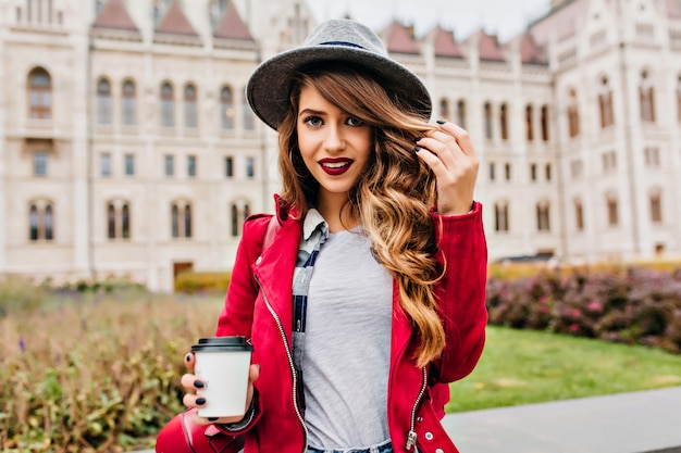 Attraente donna alla moda in posa con un sorriso dolce e tenendo la tazza di caffè al mattino