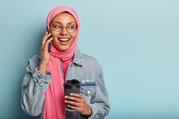 Attraente donna araba allegra ha una conversazione telefonica con un caro amico, tiene la tazza di caffè usa e getta, distoglie lo sguardo