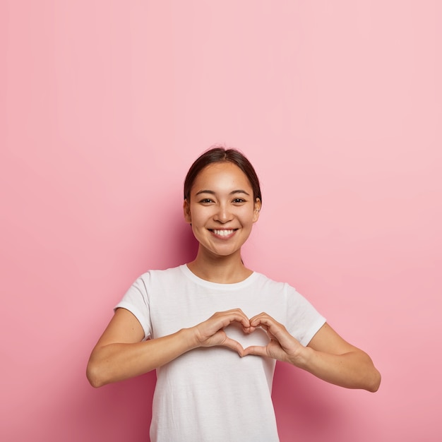 Attraente donna asiatica fa un gesto a forma di cuore, esprime amore, dice sii il mio San Valentino, sorride positivamente, indossa abiti bianchi, posa contro il muro rosa con uno spazio vuoto. Concetto di linguaggio del corpo