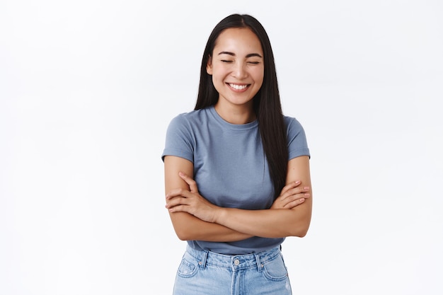 Attraente donna asiatica felice sorridente, chiudere gli occhi mentre ridacchia da uno scherzo divertente, sorridere, incrociare le mani sul petto, ridere e divertirsi, muro bianco