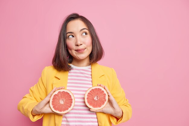 Attraente donna asiatica ha un'espressione sognante premurosa tiene metà del pompelmo mangia agrumi succosi per bruciare calorie ha una sana alimentazione posa contro il muro rosa con area spazio copia