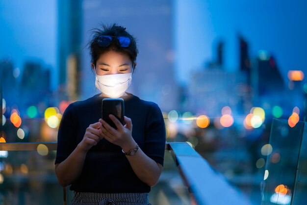 Attraente donna asiatica indossare maschera protettiva uso mano tecnologia smartphone comunicazione con sfondo di bokeh luce urbano sfondo edificio idee business concept