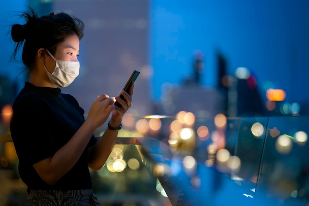 Attraente donna asiatica indossare maschera protettiva uso mano tecnologia smartphone comunicazione con sfondo di bokeh luce urbano sfondo edificio idee business concept